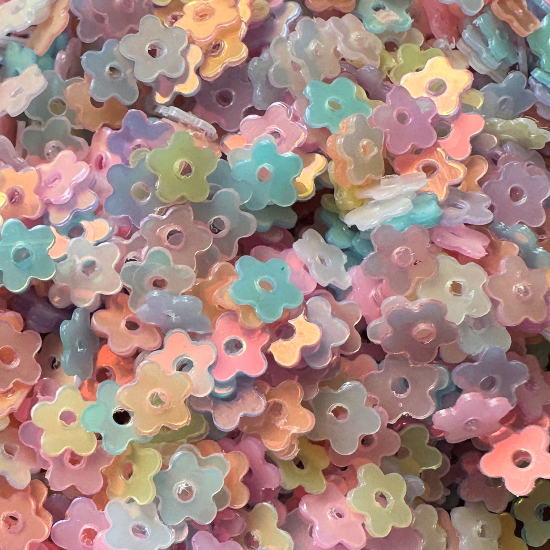 A dense pile of small, translucent flower-shaped sequins in pastel colors including pink, blue, yellow, green, and purple, each with a central hole.
